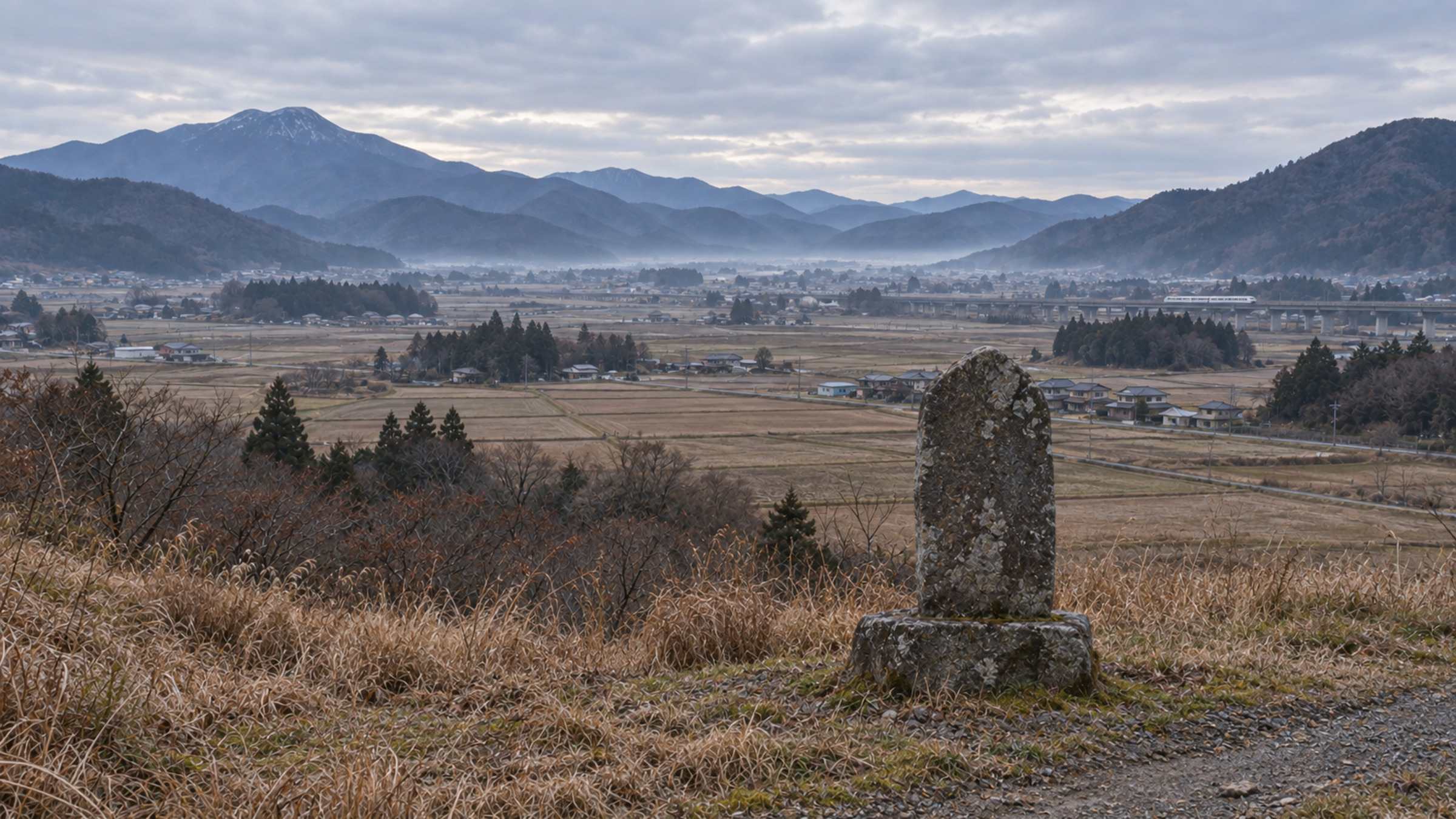 今天的同一片盆地，田地與民宅，遠處高架橋上有一列車，路邊一塊石碑背對著鏡頭